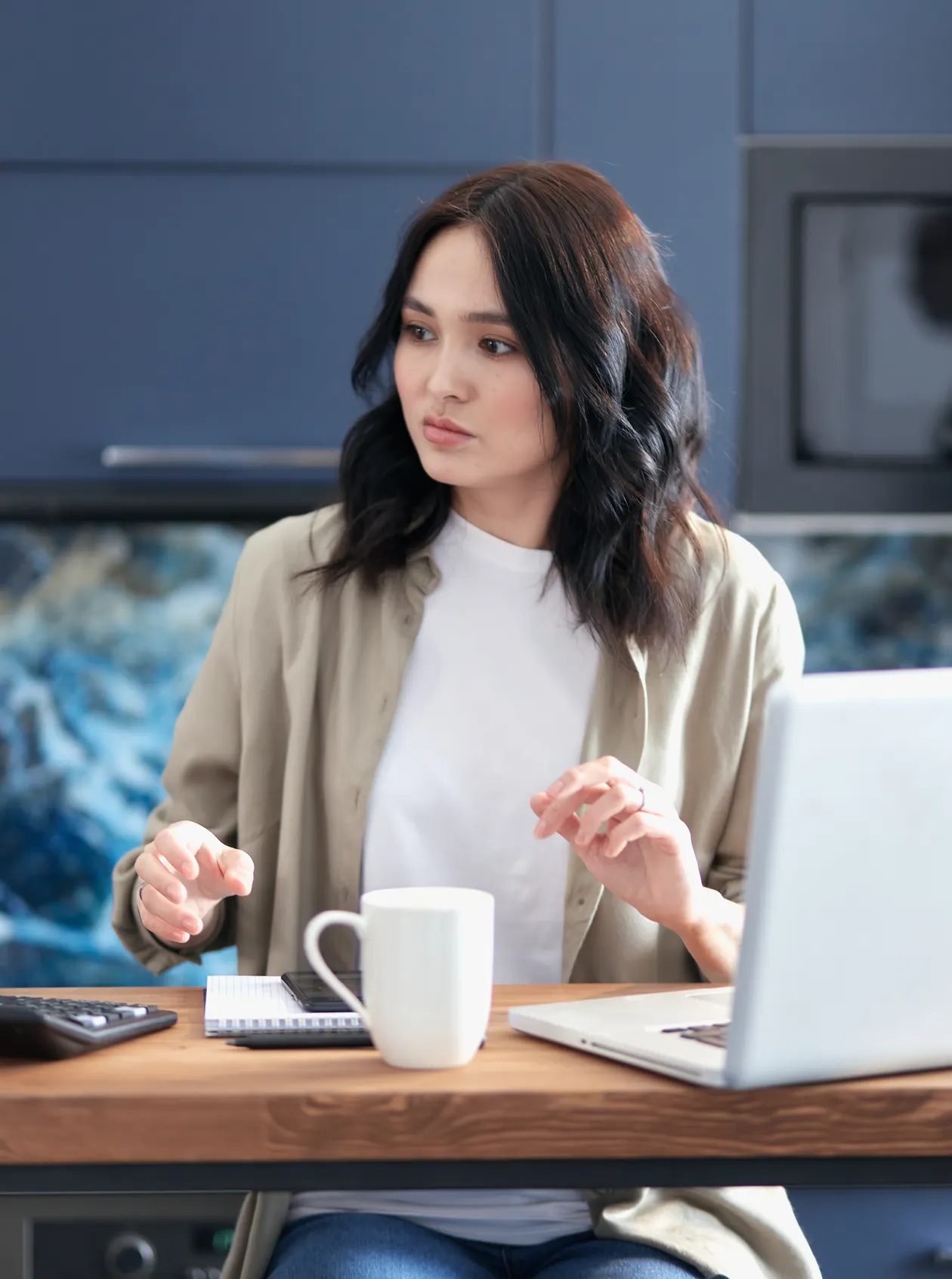 A young woman planning at a kitchen table with a laptop, calculator, notebook, and coffee